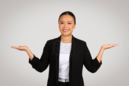 Foto de Mujer de negocios asiática balanceada con las palmas abiertas mirando hacia arriba, sonriendo con confianza, posiblemente comparando opciones o sopesando decisiones, presentadas sobre un fondo gris limpio - Imagen libre de derechos