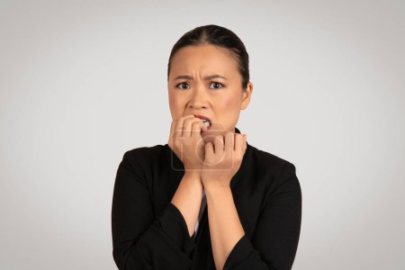 Téléchargez les photos : Femme d'affaires asiatique millénaire anxieuse dans un blazer noir avec un regard de peur, mordant nerveusement ses ongles et serrant la main, incarnant le stress ou l'inquiétude, sur fond gris - en image libre de droit