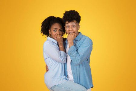 Photo for Scared young African American woman and man biting their nails, looking worried and anxious, with eyes wide open against a bright yellow background, expressing fear or anticipation - Royalty Free Image