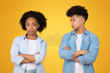 Téléchargez les photos : Jeune femme afro-américaine sceptique et homme avec les bras croisés, se donnant un regard douteux, symbolisant méfiance ou désaccord, sur un fond jaune vif - en image libre de droit