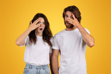Photo for Shocked and curious european couple with wide eyes covering one eye with their hands, showing expressions of surprise and intrigue, wearing white t-shirts on a yellow background - Royalty Free Image
