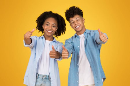Photo for Vibrant glad portrait of a young African American woman and man giving thumbs up with beaming smiles, dressed in casual light blue attire, against a sunny yellow background - Royalty Free Image