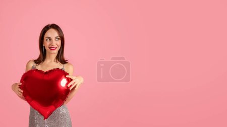 Téléchargez les photos : Une femme millénaire caucasienne avec un sourire captivant tient un ballon rouge en forme de cœur, portant une robe argentée chatoyante, symbolisant l'amour et l'affection sur un fond rose romantique - en image libre de droit