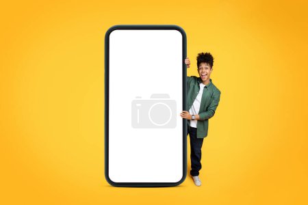 Photo for Positive stylish millennial african american guy standing by huge phone with white blank screen, showing great deal, online offer, recommending mobile app, yellow background, mockup, copy space - Royalty Free Image