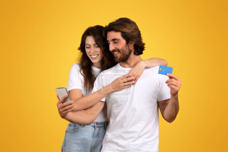 Photo for Smiling european millennial woman holding a smartphone closely while a man hugs her from behind and displays a credit card, both in white t-shirts against a yellow background - Royalty Free Image