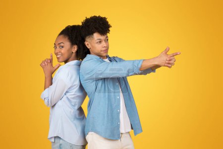 Téléchargez les photos : Deux jeunes Afro-Américains dos à dos, gesticulant ludique armes imaginaires avec leurs mains, souriant par-dessus une épaule à la caméra sur un fond jaune - en image libre de droit