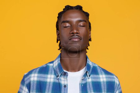 Photo for Peaceful young black man with closed eyes standing against yellow background, calm african american guy wearing checkered shirt and braided hair exuding serenity, meditating in studio, copy space - Royalty Free Image