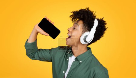 Foto de Joven y alegre chico negro cantando canciones de karaoke aisladas en el fondo amarillo del estudio, usando teléfono con pantalla vacía negra y auriculares inalámbricos. Concepto de entretenimiento - Imagen libre de derechos
