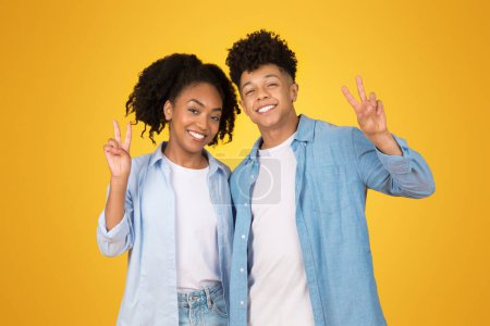Photo for Happy young African American woman and man making peace signs with their hands, both with joyful expressions, standing against a bright yellow background, symbolizing friendship and positivity - Royalty Free Image