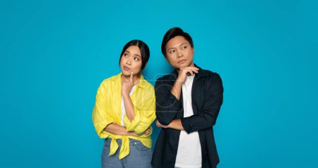 Photo for Two pensive chinese young millennial in casual clothing standing against a blue background with thoughtful expressions, seemingly pondering a decision or idea, think, brainstorm - Royalty Free Image