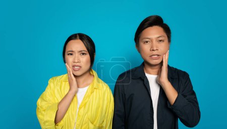 Photo for A concerned sad unhappy chinese woman and man holding their cheeks with pained expressions, possibly reacting to discomfort or a problem, against a bright blue backdrop, studio - Royalty Free Image