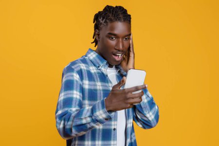 Téléchargez les photos : Offre mobile. Surpris mec noir regardant l'écran du smartphone avec excitation, homme afro-américain réagissant émotionnellement aux nouvelles en ligne, debout isolé sur fond jaune, espace de copie - en image libre de droit