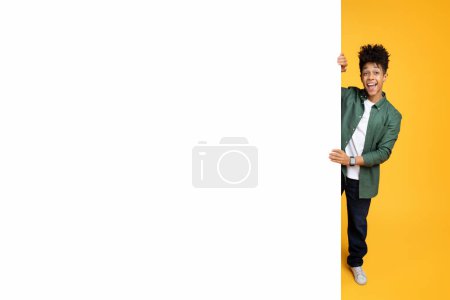 Foto de Emocionado guapo elegante millennial afroamericano chico de pie junto a blanco tablero de publicidad espacio de copia maqueta para texto o anuncio, fondo de estudio amarillo - Imagen libre de derechos