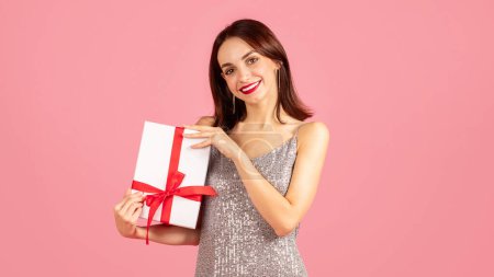 Foto de Elegante joven caucásica alegre en un vestido brillante sosteniendo una caja de regalo blanca con una cinta roja, su sonrisa expresando la alegría de dar o recibir un regalo especial - Imagen libre de derechos