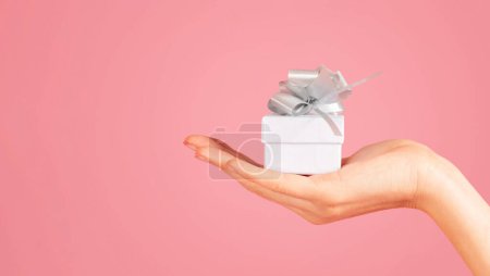 Foto de Mano abierta de persona milenaria sosteniendo una pequeña caja de regalo blanca con una cinta de plata brillante, lo que sugiere un gesto delicado y reflexivo, sobre un fondo rosa suave, recortado - Imagen libre de derechos