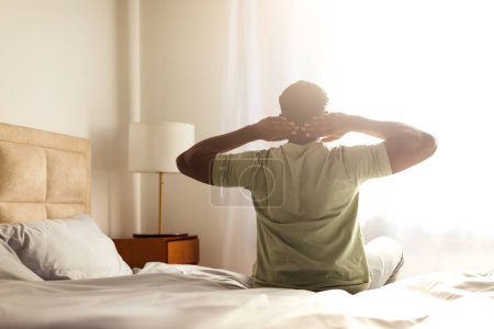 Téléchargez les photos : Homme afro-américain bien reposé en pyjama profitant d'une matinée détendue, assis sur le lit les mains derrière la tête, regardant la fenêtre et la lumière du soleil, se réveillant le matin dans la chambre d'hôtel - en image libre de droit