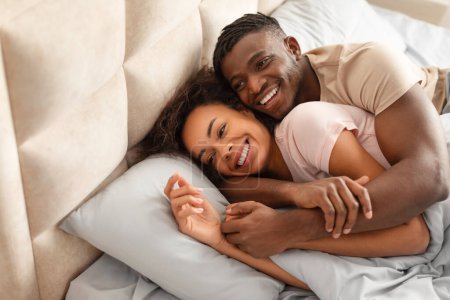 Foto de Bonita mañana. Bastante cariñosa pareja afroamericana descansando en el dormitorio juntos, abrazando y sonriendo en la cama, disfrutando del tiempo de abrazo en fin de semana, el marido abraza a la esposa desde atrás - Imagen libre de derechos