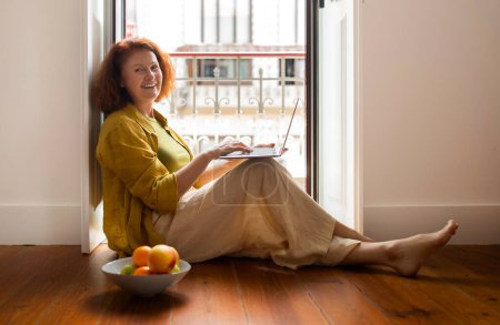 Téléchargez les photos : Femme âgée joyeuse travaillant sur son ordinateur portable près de la fenêtre à la maison, souriante femme âgée assise confortablement sur le sol par la porte ouverte du balcon, profitant d'une matinée productive, espace de copie - en image libre de droit