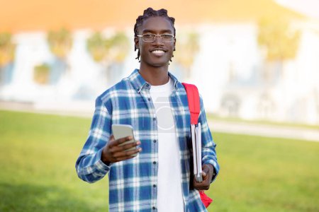 Photo for Black student guy in plaid shirt and eyeglasses uses cellphone standing outdoors, smiling to camera while showcasing the blend of technology and education for contemporary youth in university - Royalty Free Image