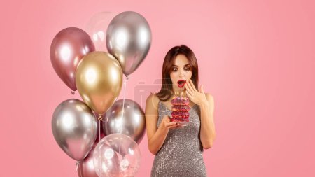 Téléchargez les photos : Étonné jeune femme caucasienne dans une robe étincelante regardant une pile de beignets colorés avec des bougies, entouré de ballons d'hélium, célébrant une occasion amusante et festive - en image libre de droit