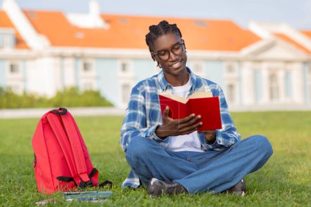 Téléchargez les photos : Heureux étudiant afro-américain aime lire des livres en plein air, portant des lunettes tout en lisant un roman assis sur la pelouse au parc du campus universitaire en plein air. Concept de loisirs et d'apprentissage - en image libre de droit