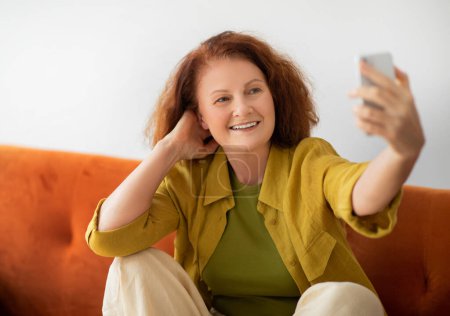 Téléchargez les photos : Joyeuse femme âgée avec smartphone prenant selfie à la maison, belle femme âgée posant à la caméra tout en se relaxant sur le canapé dans le salon, mettant en valeur l'engagement technologique, espace libre - en image libre de droit