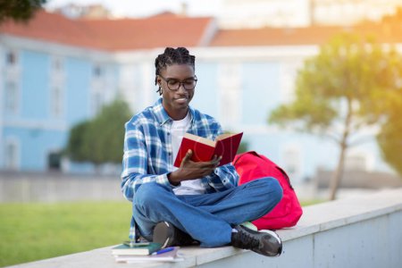 Foto de Estudiante joven negro feliz en anteojos leyendo un libro al aire libre, disfrutando estudiando literatura en el campus universitario Park al aire libre, sentado con mochila interactuando con una nueva novela - Imagen libre de derechos