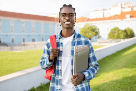 Téléchargez les photos : Un étudiant noir joyeux tenant des cahiers d'exercices, un ordinateur et un sac à dos tout en posant contre le bâtiment du campus, souriant à la caméra, prêt à étudier à l'étranger. Mode de vie de stagiaire - en image libre de droit