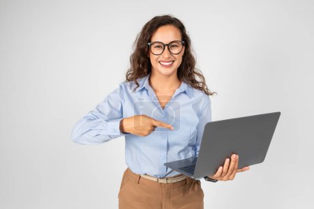 Téléchargez les photos : Une jeune femme professionnelle joyeuse portant des lunettes, une chemise bleue et des points de pantalon beige sur un écran d'ordinateur portable qu'elle tient, indiquant un point d'intérêt ou de présentation - en image libre de droit