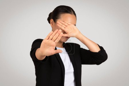 Téléchargez les photos : Femme d'affaires asiatique professionnelle en costume noir fait un geste d'arrêt avec ses mains, couvrant ses yeux, indiquant une obstruction visuelle ou d'évitement, contre un fond gris - en image libre de droit