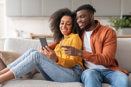Téléchargez les photos : Joyeux jeune couple noir s'amuser avec téléphone portable websurfing tout en se prélassant confortablement sur le canapé, homme pointant vers le téléphone défilement contenu Internet intéressant, à l'intérieur de la maison salon - en image libre de droit