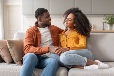 Téléchargez les photos : Joyeux couple d'amoureux noirs millénaires profitant du temps ensemble à la maison, heureux homme et femme afro-américain embrasser et parler assis sur le canapé dans le salon moderne. Amour et relation - en image libre de droit