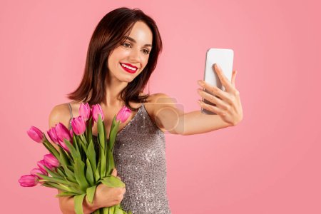 Photo for Smiling millennial caucasian woman takes a selfie with her smartphone, capturing the moment with a bunch of pink tulips in hand, wearing a sequined dress on a pink background - Royalty Free Image