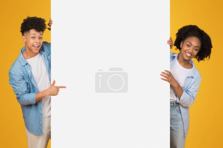 Téléchargez les photos : Adolescents afro-américains excités tenant et pointant vers une bannière blanche vierge avec de grands sourires, sur un fond jaune vif, idéal pour les publicités et les annonces - en image libre de droit