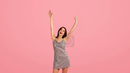 Überschwänglich fröhliche kaukasische junge Frau in einem glitzernden silbernen Kleid hebt freudig die Hände in die Luft, genießt Party, Spaß, Tanz, Feiern auf rosa Hintergrund, Panorama