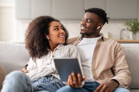 Foto de Pareja joven afroamericana relajada que lleva auriculares escuchando música juntos mientras descansan en el sofá en la sala de estar moderna, abrazándose y sonriéndose mutuamente disfrutando de la canción en casa - Imagen libre de derechos