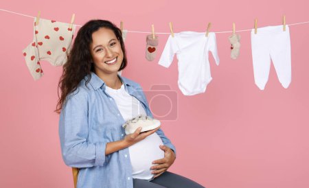 Photo for Radiant expecting mother in denim shirt cradles a baby shoe, with a clothesline of white and heart-adorned garments behind her on a soft pink backdrop, hugging her big tummy, copy space - Royalty Free Image