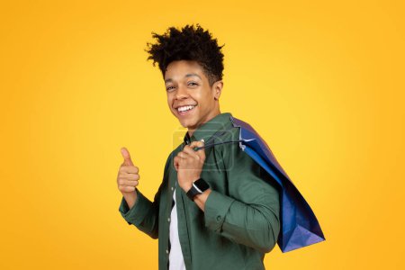 Foto de Cool guapo millennial afroamericano chico con bolsa de compras en la mano mostrando el pulgar hacia arriba y sonriendo a la cámara, aislado en el fondo del estudio amarillo. Venta temporada, consumismo - Imagen libre de derechos