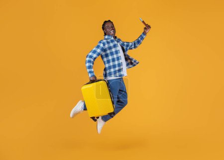Téléchargez les photos : Portrait d'un jeune homme noir heureux sautant avec une valise et un passeport, prêt pour un voyage de vacances, homme afro-américain excité courant avec des bagages sur fond jaune, prise de vue pleine longueur, espace de copie - en image libre de droit