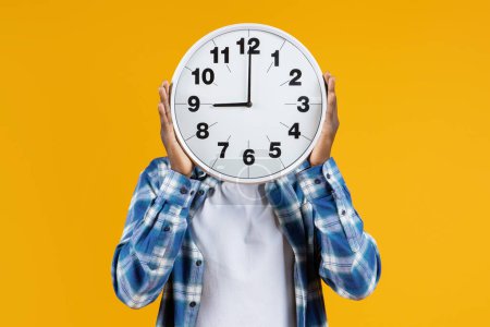 Photo for Unrecognizable black male holding large round clock in front of his face, african american man in plaid shirt standing on yellow background, representing concept of time management and punctuality - Royalty Free Image