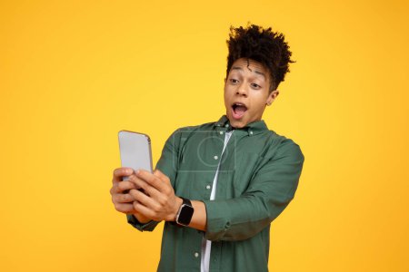 Téléchargez les photos : Incroyable beau millénariste élégant afro-américain gars regardant l'écran de smartphone dans ses mains et grimaçant, lire des nouvelles choquantes, contenu à couper le souffle, fond jaune - en image libre de droit