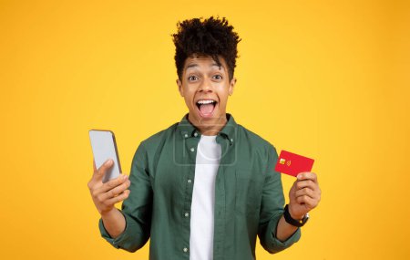 Téléchargez les photos : Joyeux beau jeune homme noir avec carte de crédit bancaire en plastique et smartphone dans ses mains posant isolé sur fond jaune. Achats en ligne, banque, e-commerce, cashback - en image libre de droit