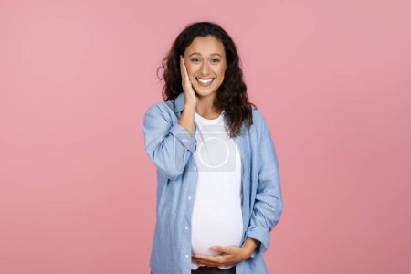 Téléchargez les photos : Plan studio de femme enceinte heureuse touchant son visage et son ventre et souriant, posant sur fond de studio rose, femme enceinte souriante jouissant d'un temps de grossesse sain - en image libre de droit