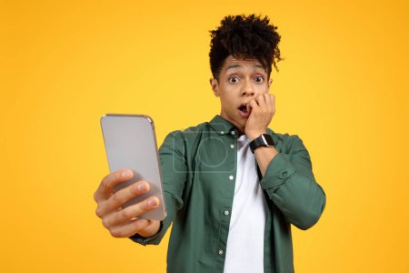 Téléchargez les photos : Choqué beau mec élégant millénariste afro-américain tenant smartphone dans sa main et grimaçant, lire des nouvelles choquantes, contenu à couper le souffle, fond jaune, espace de copie - en image libre de droit