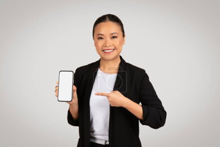 Téléchargez les photos : Femme d'affaires asiatique souriante présentant un smartphone avec un écran vide, le pointant d'une main, suggérant une application ou une fonctionnalité, idéal pour le marketing lié à la technologie - en image libre de droit