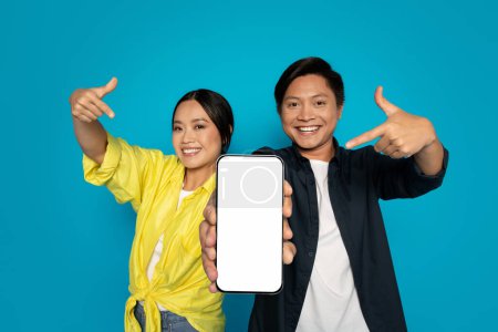Téléchargez les photos : Enthousiaste heureux chinois femme et homme pointant avec excitation vers un smartphone avec un écran blanc, présentant une nouvelle application ou une publicité sur un fond turquoise, studio - en image libre de droit