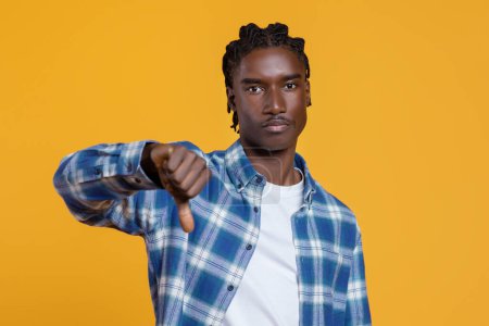 Photo for Dissatisfied young black man showing thumb down gesture at camera, serious african american guy wearing plaid shirt standing isolated against yellow studio background, expressing dislike, copy space - Royalty Free Image