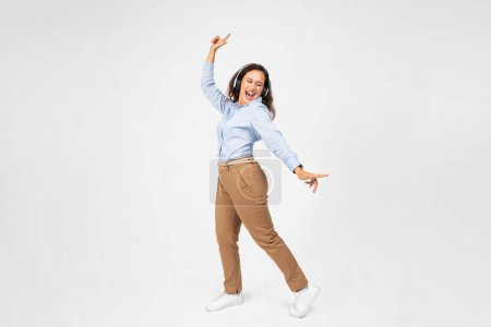 Foto de Una animada joven europea con el pelo rizado, vestida con una camisa azul y pantalones marrones, baila alegremente con los auriculares puestos, expresando libertad y felicidad en su movimiento - Imagen libre de derechos