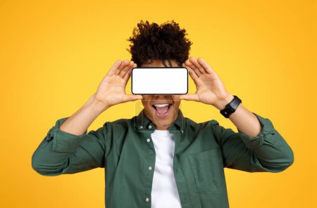 Téléchargez les photos : Black guy holding smartphone with white blank screen over his eyes and grimacing, mockup copy space for online offer deal advertising, application mobile, fond jaune studio - en image libre de droit