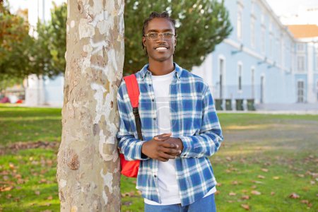 Téléchargez les photos : Étudiant afro-américain confiant avec sac à dos portant des lunettes, se tient dans le parc du campus universitaire ensoleillé, regardant la caméra avec un sourire joyeux. Apprendre en plein air académique - en image libre de droit